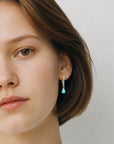 Modelo luciendo aretes huggies Queens Candy de plata 925 con baño dorado y dije de gota turquesa sintética.